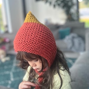 May include: A child wearing a red and yellow knitted hat with a pointed top. The hat has a long tie that hangs down the back of the child's neck.