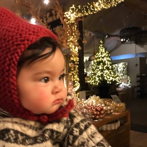 May include: A baby wearing a red knitted hat with a white and black patterned sweater. The baby is looking to the right of the frame. The background is blurry and shows a Christmas tree with lights.