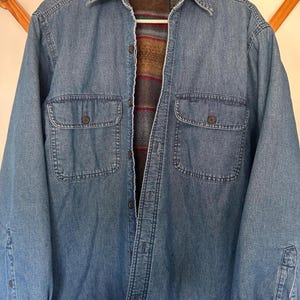 May include: A blue denim shirt jacket with two chest pockets and button closures. The jacket has a brown and red striped lining. The sleeves have buttoned cuffs. The jacket is hanging on a wooden rack.