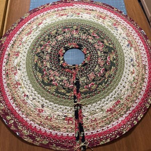 May include: A circular quilted tree skirt featuring concentric rings of fabric in shades of red, green, black, and cream. The design includes floral and botanical patterns with decorative stitching throughout. It has a center opening and three ties.