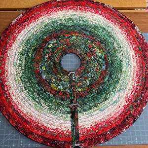 May include: A circular fabric tree skirt featuring a coiled rope construction with a gradient of red, white, and green patterned cotton fabrics. The design includes a central circular cutout and a straight slit with three small grey fabric ties.