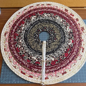May include: A circular fabric tree skirt featuring concentric rings of quilted fabric in black, deep red, and cream. The design includes intricate patterns, gold accents, and script text. A vertical fabric strip with three twine ties extends from the center.