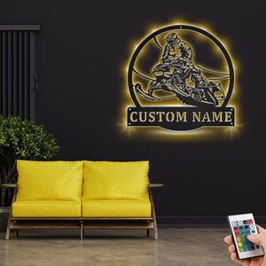 May include: A black metal silhouette of a person riding a snowmobile in a circle with a gold 'CUSTOM NAME' sign below. The silhouette is illuminated with warm white LED lights.