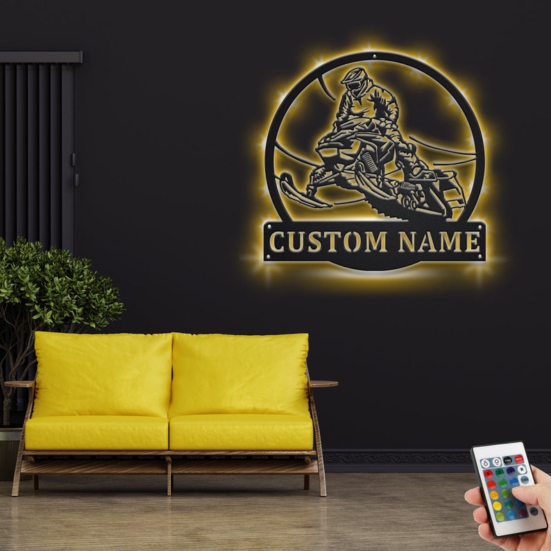 May include: A black metal silhouette of a person riding a snowmobile in a circle with a gold 'CUSTOM NAME' sign below. The silhouette is illuminated with warm white LED lights.