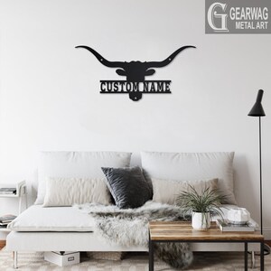 May include: Black metal silhouette of a longhorn steer head with large horns and a nameplate that reads "CUSTOM NAME". The silhouette is mounted on a white wall above a white couch with pillows and a throw blanket.