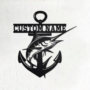 Pode incluir: Arte de parede de metal preto com uma âncora com um marlin saltando para fora da água. A âncora tem uma faixa na parte superior que diz "CUSTOM NAME".