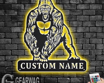 May include: A black metal silhouette of a gorilla with a yellow glow. The gorilla is standing on a black metal sign with the text "CUSTOM NAME" in white. The sign is illuminated with a yellow glow. A remote control for LED lights is visible in the bottom right corner of the image.