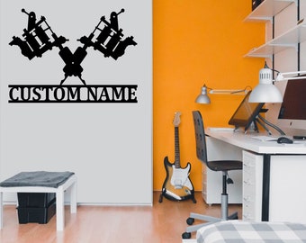 May include: Black silhouette of two tattoo machines crossed with the text "CUSTOM NAME" below. The image is on a white wall in a room with a guitar, desk, and chair.