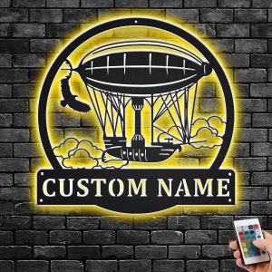 Puede incluir: Un letrero de metal negro con una silueta de un dirigible con nubes y el texto "CUSTOM NAME". El letrero está iluminado con un brillo amarillo.