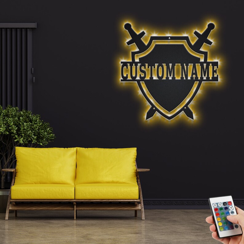 May include: A black metal shield with two swords crossed behind it. The shield has a yellow outline and the words "CUSTOM NAME" written across the center. The shield is mounted on a black wall with a yellow couch in the foreground.