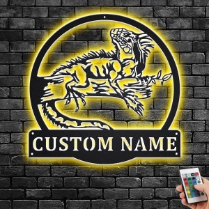 Puede incluir: Silueta de iguana de metal negro con un brillo amarillo, montada en una pared de ladrillo. La iguana está posada en una rama con su cola enroscada alrededor de la rama. El letrero de metal tiene espacio para un nombre personalizado, que dice "CUSTOM NAME". Un control remoto para cambiar el color de la luz es visible en la esquina inferior derecha.