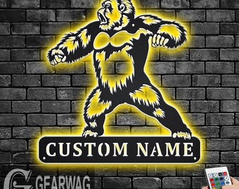 May include: A black metal silhouette of a gorilla with a yellow glow. The gorilla is standing with its arms outstretched. Below the gorilla is a black metal sign with the text "CUSTOM NAME" in white. The sign is also lit with a yellow glow. The image is on a brick wall background.