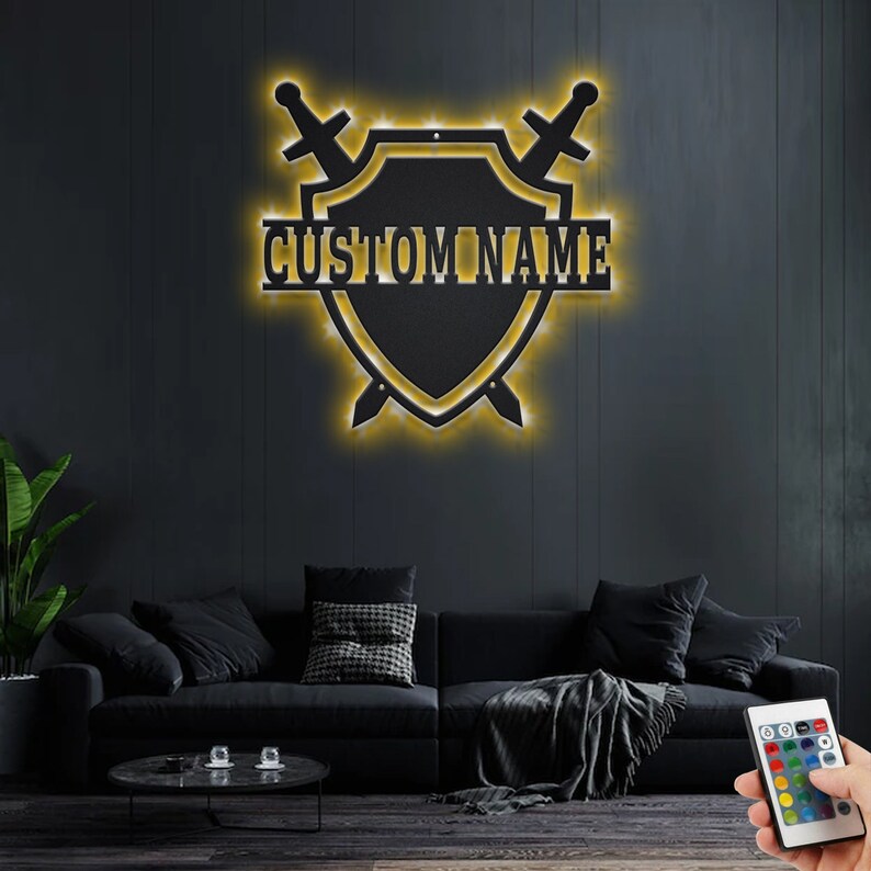 May include: A black metal shield with two swords crossed behind it. The shield has a yellow outline and the words "CUSTOM NAME" are written across the center. The shield is illuminated with warm white LED lights.