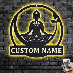 K&ouml;nnte beinhalten: Eine schwarze Wanddekoration aus Metall mit einer Silhouette einer Person, die in einem Kreis mit gelbem Schein meditiert. Die Worte "CUSTOM NAME" befinden sich am unteren Rand des Kreises.