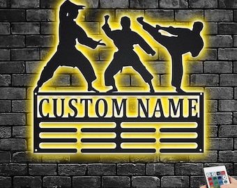 May include: Black metal wall art featuring three karate figures in action poses, illuminated by a yellow glow. The sign includes the text "CUSTOM NAME" and a series of horizontal lines. The background is a dark brick wall.