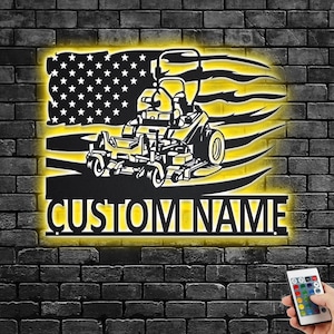 Puede incluir: Una silueta negra de metal de un cortacésped de conducción con una bandera estadounidense detrás. El cortacésped está iluminado en amarillo. El texto "CUSTOM NAME" está debajo del cortacésped.