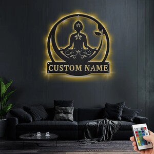K&ouml;nnte beinhalten: Eine schwarze Wanddekoration aus Metall mit einem runden Design, das eine Silhouette einer Person zeigt, die im Lotussitz meditiert. Das Design enth&auml;lt florale Akzente und ein anpassbares Namensschild mit der Aufschrift "CUSTOM NAME". Die Dekoration wird mit warmwei&szlig;en LED-Lichtern beleuchtet.