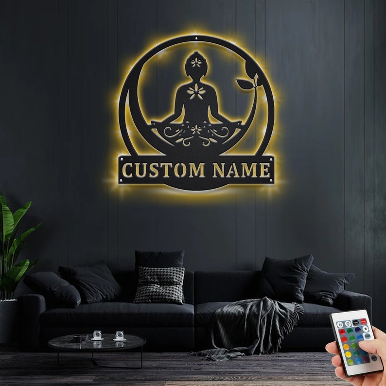 K&ouml;nnte beinhalten: Eine schwarze Wanddekoration aus Metall mit einem runden Design, das eine Silhouette einer Person zeigt, die im Lotussitz meditiert. Das Design enth&auml;lt florale Akzente und ein anpassbares Namensschild mit der Aufschrift "CUSTOM NAME". Die Dekoration wird mit warmwei&szlig;en LED-Lichtern beleuchtet.