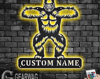 May include: A black metal silhouette of a gorilla with yellow LED lights behind it. The gorilla is standing with its arms outstretched. Below the gorilla is a black metal sign with the text "CUSTOM NAME" in white LED lights. A remote control for the lights is visible in the bottom right corner of the image.