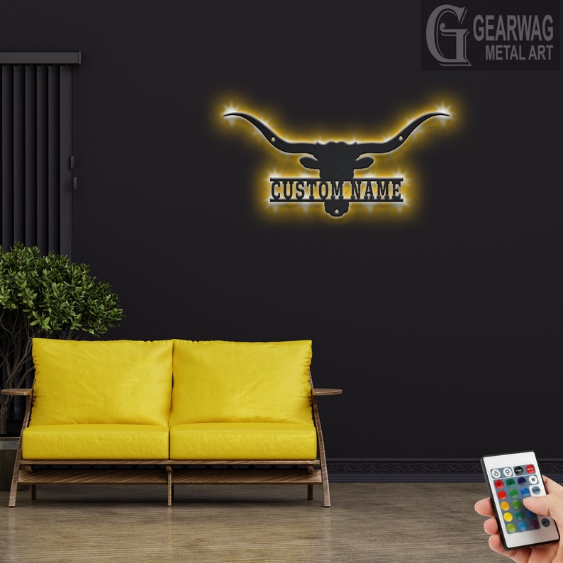May include: A black metal longhorn steer head silhouette with a custom nameplate and LED lights. The lights are illuminated in a warm yellow color. The steer head is mounted on a black wall. A yellow couch is in the foreground. A remote control for the lights is in the lower right corner of the image.