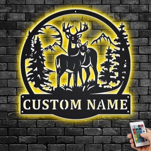 Op de afbeelding: Een zwart metalen silhouet van twee herten die in een bos scène staan met een gele gloed rond de randen. Het silhouet is gemonteerd op een bakstenen muur met een zwarte metalen plaat eronder met de tekst "CUSTOM NAME".