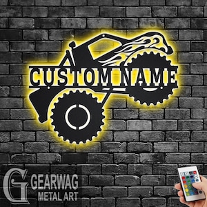 Op de afbeelding: Zwarte metalen silhouet van een monstertruck met vlammen, met de woorden "CUSTOM NAME" in het midden. De truck is verlicht met gele LED-verlichting.