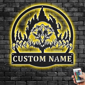 May include: Black metal wall art featuring an eagle with its claws outstretched, emerging from a fiery background. The art is illuminated with yellow LED lights and includes a customizable nameplate that reads "CUSTOM NAME".
