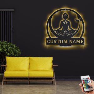 K&ouml;nnte beinhalten: Schwarze Wanddekoration aus Metall mit einer Silhouette einer Person in einer Yoga-Pose, umgeben von floralen Designs. Die Dekoration ist mit warmwei&szlig;en LED-Lichtern beleuchtet und hat ein anpassbares Namensschild mit der Aufschrift "CUSTOM NAME".