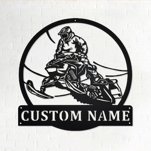 May include: Black metal silhouette of a person riding a snowmobile in a circle with the words "CUSTOM NAME" below.