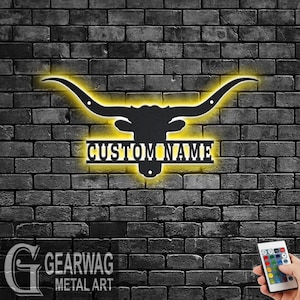 May include: Black metal silhouette of a longhorn steer head with large horns, illuminated with yellow light. The name "CUSTOM NAME" is displayed on a metal plaque below the steer head.