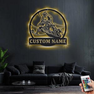 May include: A black metal wall art piece featuring a silhouette of a person riding a snowmobile. The snowmobile is surrounded by a circle of lights. The words "CUSTOM NAME" are displayed below the snowmobile.