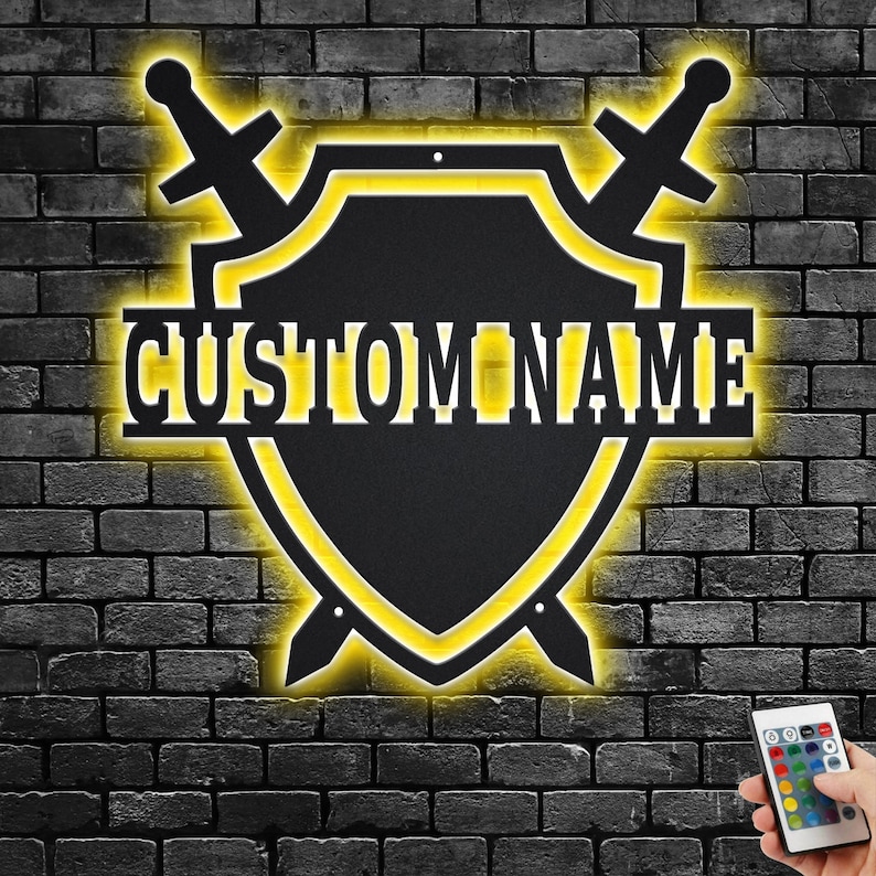 May include: A black metal shield with two swords crossed behind it. The shield has a yellow outline and the words "CUSTOM NAME" are printed on the shield. The shield is mounted on a brick wall.