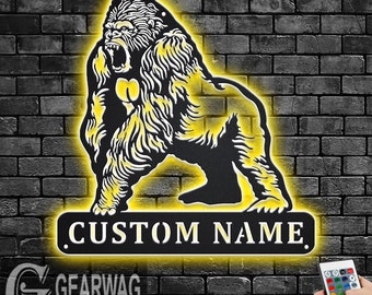 May include: A black metal silhouette of a gorilla with a yellow glow around it. The gorilla is standing on a black metal plaque with the text "CUSTOM NAME" in white. The plaque is illuminated with a yellow glow. A remote control for LED lights is visible in the bottom right corner of the image.