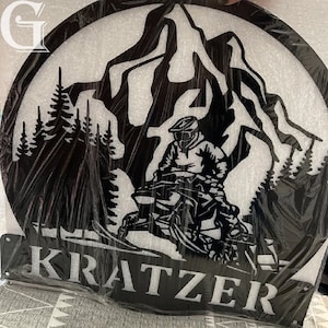 May include: Black metal silhouette of a person riding a snowmobile through a mountain landscape with trees. The name "KRATZER" is displayed below the image.