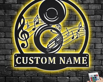 Puede incluir: Arte de pared de metal negro con una tuba con notas musicales y una placa de nombre personalizable. La placa de nombre dice "CUSTOM NAME". El arte está iluminado con una luz amarilla cálida.