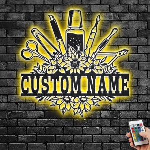 May include: A black metal sign with a floral design and the text "CUSTOM NAME". The sign is illuminated with yellow lights and features various nail salon tools, including nail clippers, a nail file, and a bottle of nail polish.