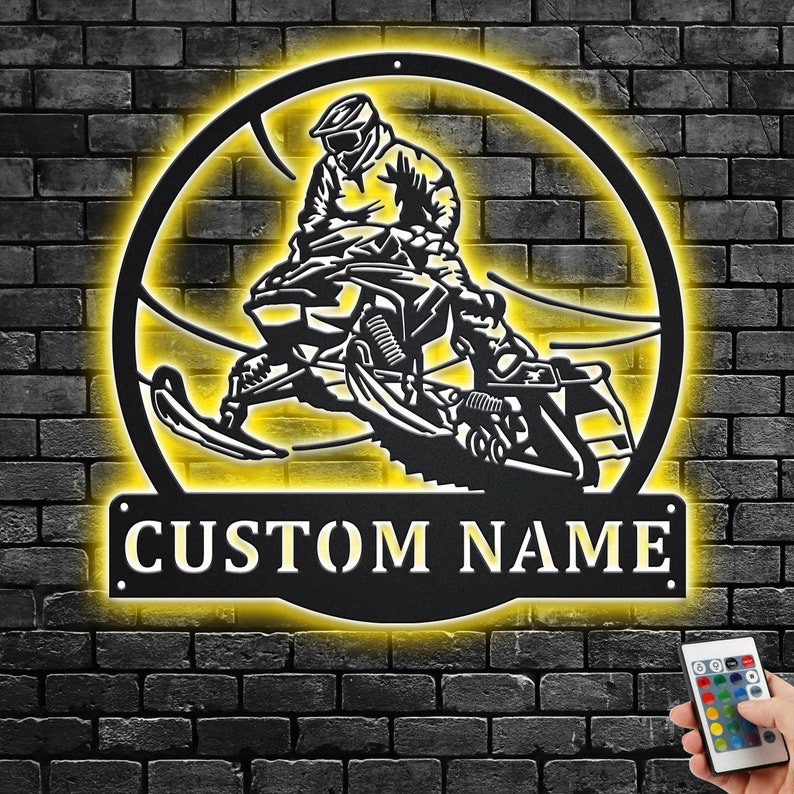 May include: Black metal silhouette of a person riding a snowmobile with a yellow glow around it. The words "CUSTOM NAME" are below the snowmobile.