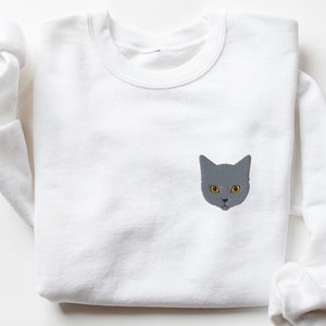 May include: White sweatshirt with a gray cat face embroidered on the front. The cat has yellow eyes.