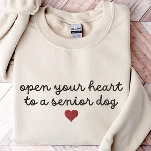 May include: A beige sweatshirt with the text "open your heart to a senior dog" and a red heart printed on the front.