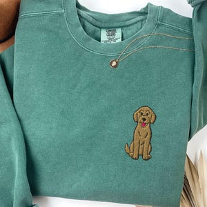 May include: Light green sweatshirt with an embroidered brown dog on the front. The sweatshirt has a crew neck and long sleeves. A gold necklace with a small pendant is visible. The text "LIGHT GREEN" is at the bottom.