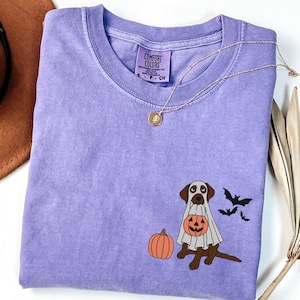 May include: A purple t-shirt with a graphic of a brown dog dressed as a ghost holding a pumpkin. The dog is wearing a white ghost costume with a jack-o-lantern face. There are two black bats flying above the dog. The shirt has a tag that says "Comfort Colors".