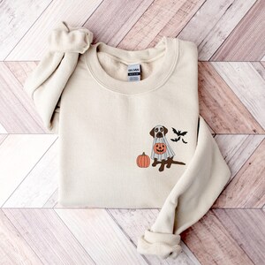May include: A cream-coloured sweatshirt featuring a graphic of a brown dog dressed as a ghost, holding a jack-o'-lantern. Two bats are flying above the dog, and a pumpkin is on the ground.