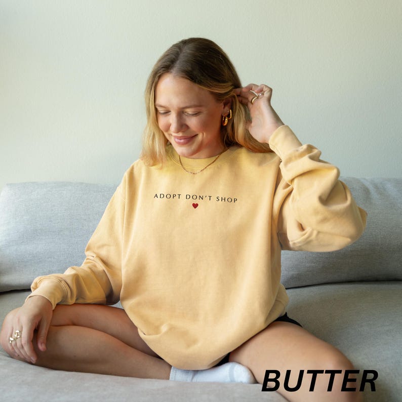 May include: A pale yellow sweatshirt featuring the text "ADOPT DON'T SHOP" above a small red heart. The word "BUTTER" is printed in a black font at the bottom of the image. The sweatshirt has long sleeves.