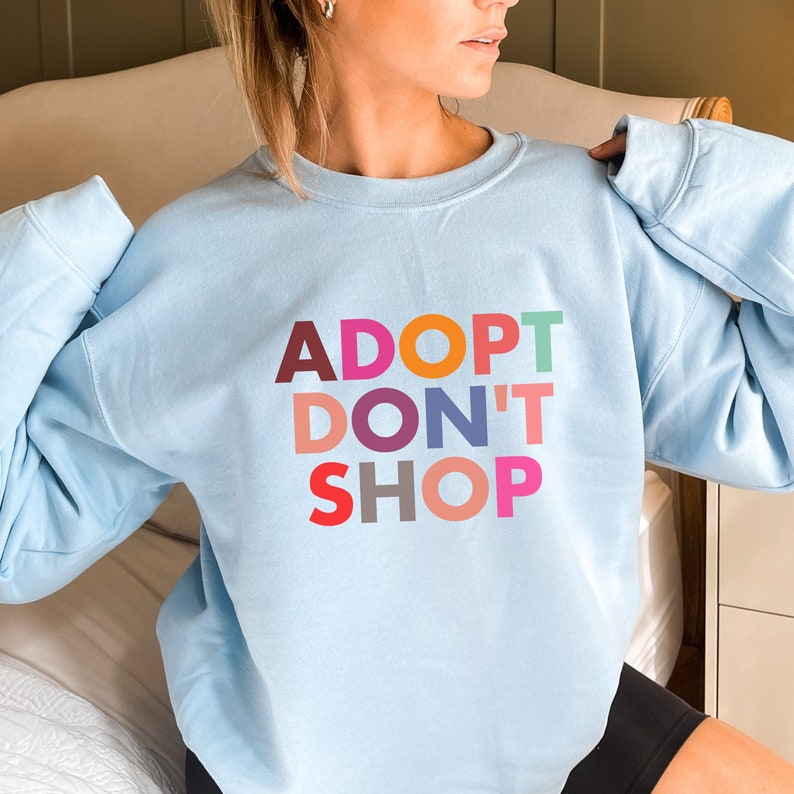 May include: A light blue sweatshirt with the text "ADOPT DON'T SHOP" in colorful letters. The letters are in different colors, including red, orange, yellow, green, blue, purple, and pink.