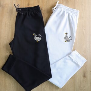 May include: Two pairs of sweatpants, one black and one white, with a gray goose and the word "silly" embroidered on the left leg.