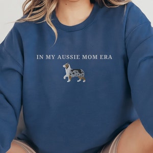 以下が含まれることがあります： 「IN MY AUSSIE MOM ERA」という文字と、オーストラリア・シェパードの刺繍が施されたネイビーブルーのスウェットシャツです。