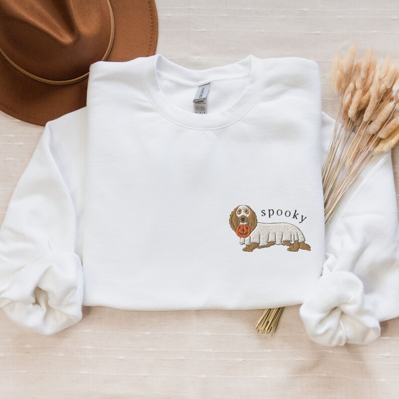 May include: White sweatshirt with a brown and white dog wearing a ghost costume and holding a jack-o-lantern. The text "spooky" is above the dog.