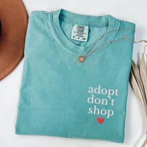 May include: Teal blue t-shirt with the text "adopt don't shop" in white with a red heart underneath. The shirt has a tag that says "Comfort Colors" and a gold necklace with a charm is draped over the shirt.