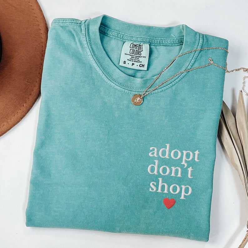May include: Teal blue t-shirt with the text "adopt don't shop" in white with a red heart underneath. The shirt has a tag that says "Comfort Colors" and a gold necklace with a charm is draped over the shirt.