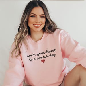 May include: A pink sweatshirt with the text "open your heart to a senior dog" in black lettering and a small red heart.